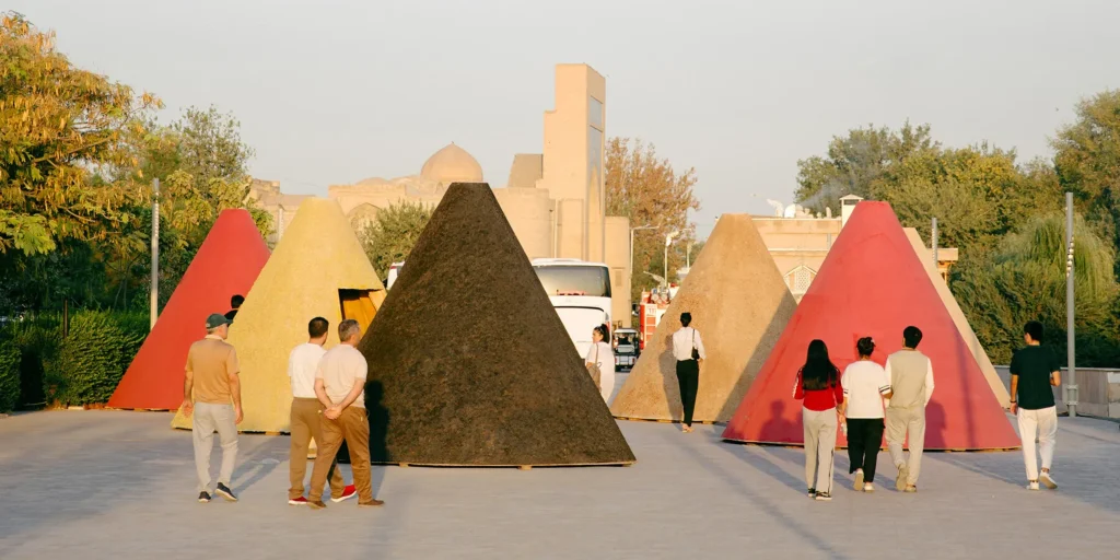 Group of visitors walking among large colourful conical kiosks designed by Ariel André in red, yellow, black and pink, set in an outdoor plaza with historic buildings in the background at dusk