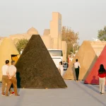 Group of visitors walking among large colourful conical kiosks in red, yellow, black and pink, set in an outdoor plaza with historic buildings in the background at dusk