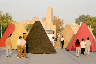 Group of visitors walking among large colourful conical kiosks in red, yellow, black and pink, set in an outdoor plaza with historic buildings in the background at dusk
