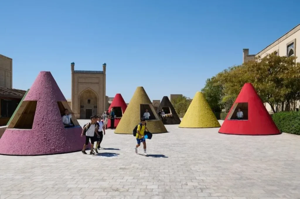 Colourful conical kiosks in pink, red, yellow and black arranged in a sunny historic plaza, with children and visitors walking between them under a clear blue sky