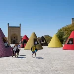 Colourful conical kiosks in pink, red, yellow and black arranged in a sunny historic plaza, with children and visitors walking between them under a clear blue sky