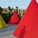 Large bright red conical structures standing in a paved courtyard, with a person’s shadow cast on the nearest cone and historic buildings in the background