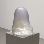 Translucent pearlescent glass form with an organic, rounded shape resting on a grey concrete block against a plain white background