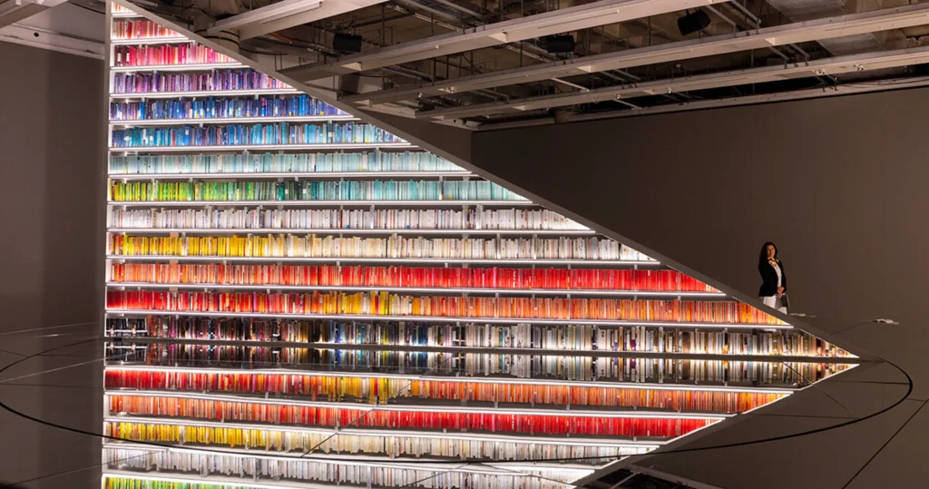 A large, illuminated bookshelf filled with color-coordinated books arranged in a spectrum, reflected on a polished floor while artist Es Devlin stands in the background.