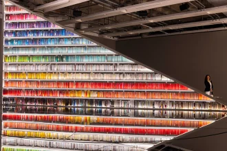 A large, illuminated bookshelf filled with color-coordinated books arranged in a spectrum, reflected on a polished floor while artist Es Devlin stands in the background.