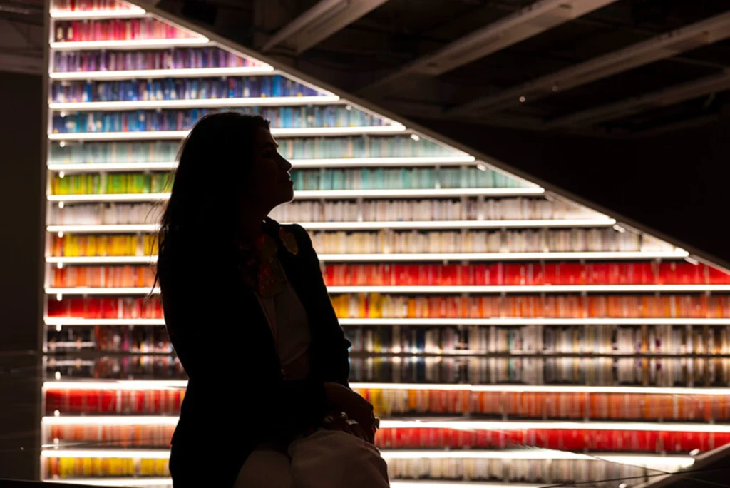 Silhouette of Es Devlin sitting in front of a brightly lit, triangular bookshelf filled with colorful books.