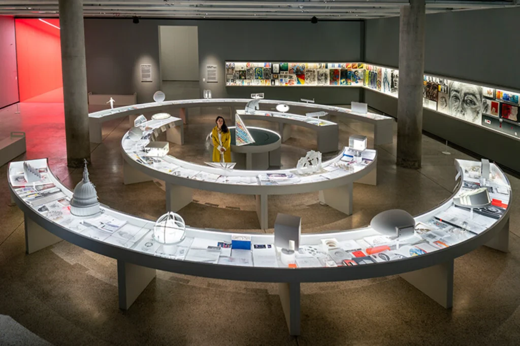 Wide high-angle view of a gallery space featuring a large spiral display table filled with models, sketches, and design artifacts with Es Devlin standing near the center.