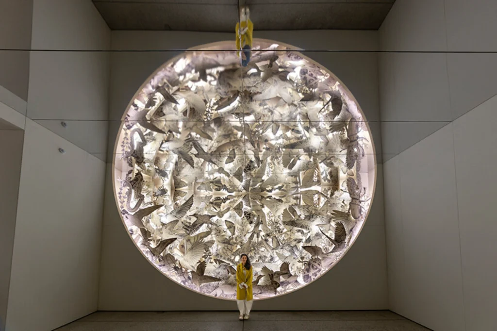 Es Devlin standing in a bright yellow coat before a large circular wall installation featuring intricate, paper-cut depictions of animals and birds.