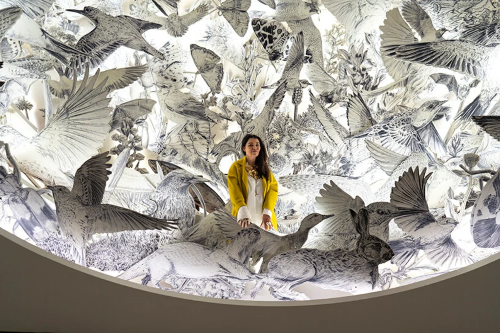 Close-up shot of Es Devlin surrounded by a dense, circular paper installation filled with detailed drawings of flying birds, insects, and mammals.