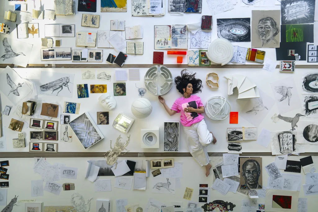 High-angle overhead shot of Es Devlin lying on a wall layout surrounded by various sketches, 3D architectural models, photos, and notes.