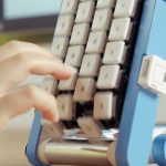 Hand pressing keys on the blue conveyor-belt keyboard as the keys move past the fingers