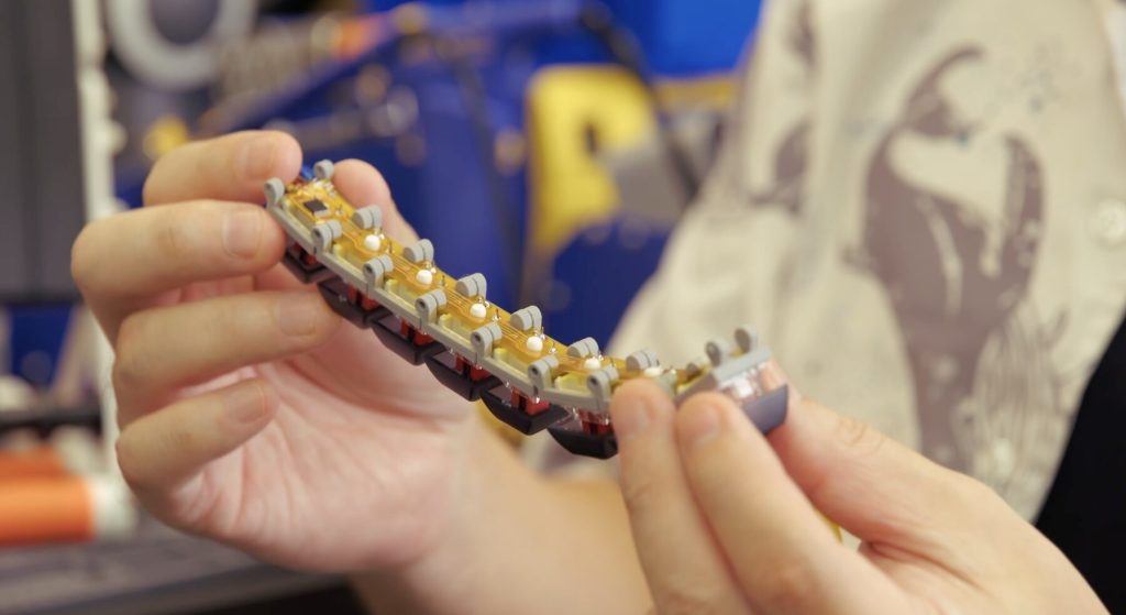 Close-up of hands holding a flexible circuit strip with small rollers and electronic components