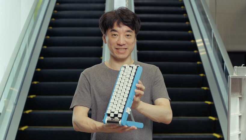 Smiling man holding the blue Gboard conveyor-belt keyboard in front of an escalator