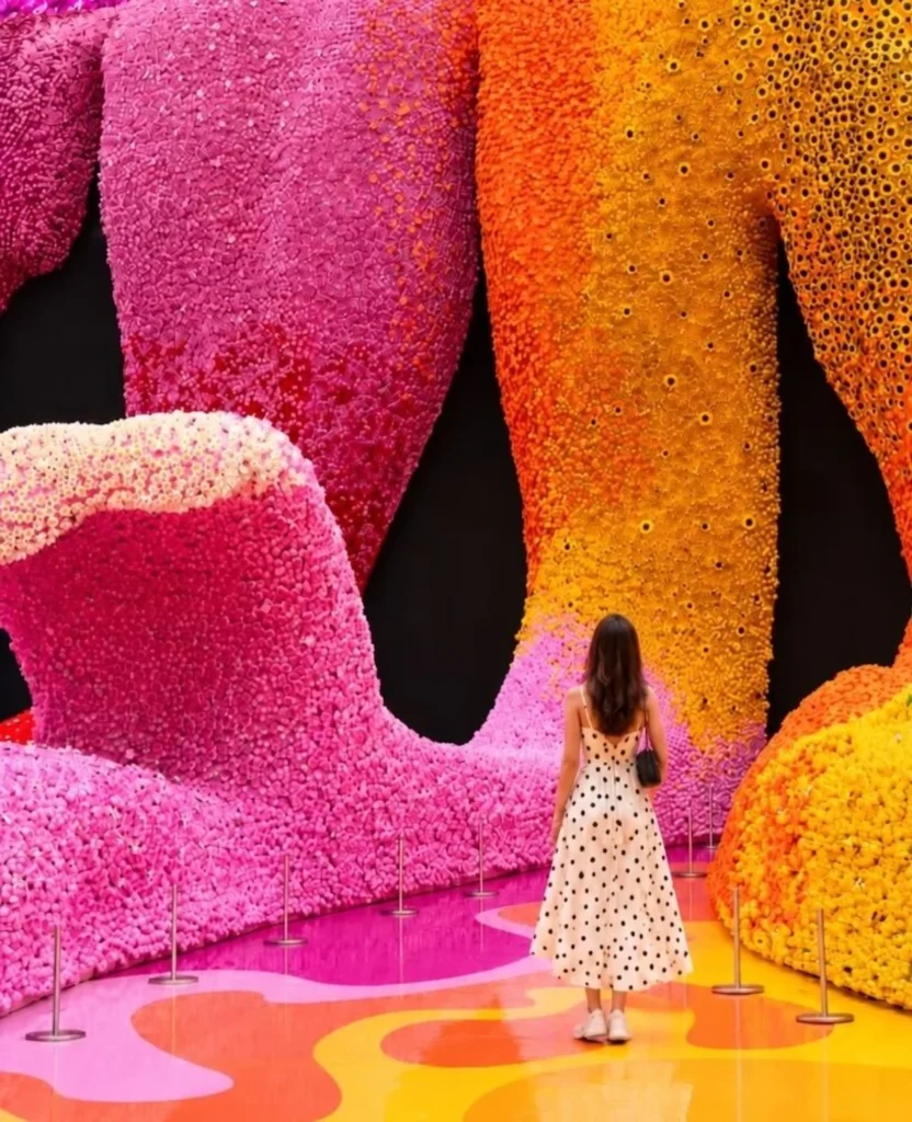 Woman in a polka-dot dress standing before towering pink and orange LEGO flower walls inside the exhibition space