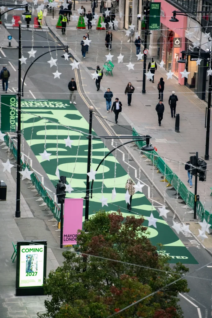 Elevated view of Oxford Street's pedestrianization trial in central London, featuring large green road graphics, temporary barriers, pedestrians, and festive street decorations