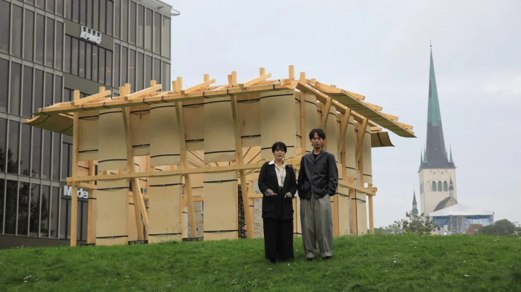 Two people standing in front of the wooden Resonance Pavilion with a church spire and modern building in the background