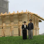 Two people standing in front of the wooden Resonance Pavilion with a church spire and modern building in the background