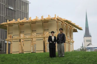 Two people standing in front of the wooden Resonance Pavilion with a church spire and modern building in the background