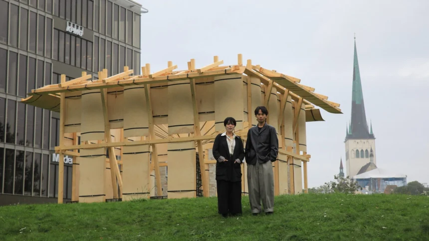 Two people standing in front of the wooden Resonance Pavilion with a church spire and modern building in the background