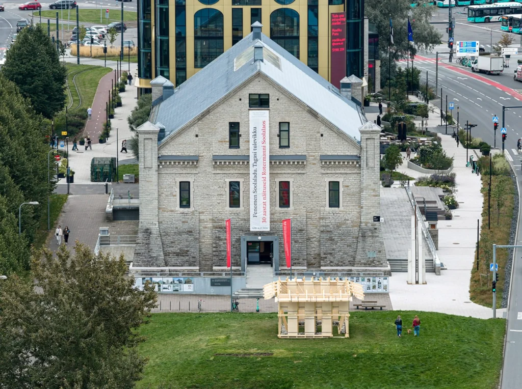 Aerial view of the wooden Resonance Pavilion sitting on a grassy traffic island in front of the Estonian Museum of Architecture
