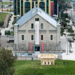 Aerial view of the wooden Resonance Pavilion sitting on a grassy traffic island in front of the Estonian Museum of Architecture