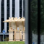 Side view of the Resonance Pavilion seen through a window, with its light timber frame and plywood panels against a modern building