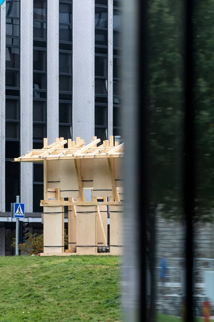 Side view of the Resonance Pavilion seen through a window, with its light timber frame and plywood panels against a modern building