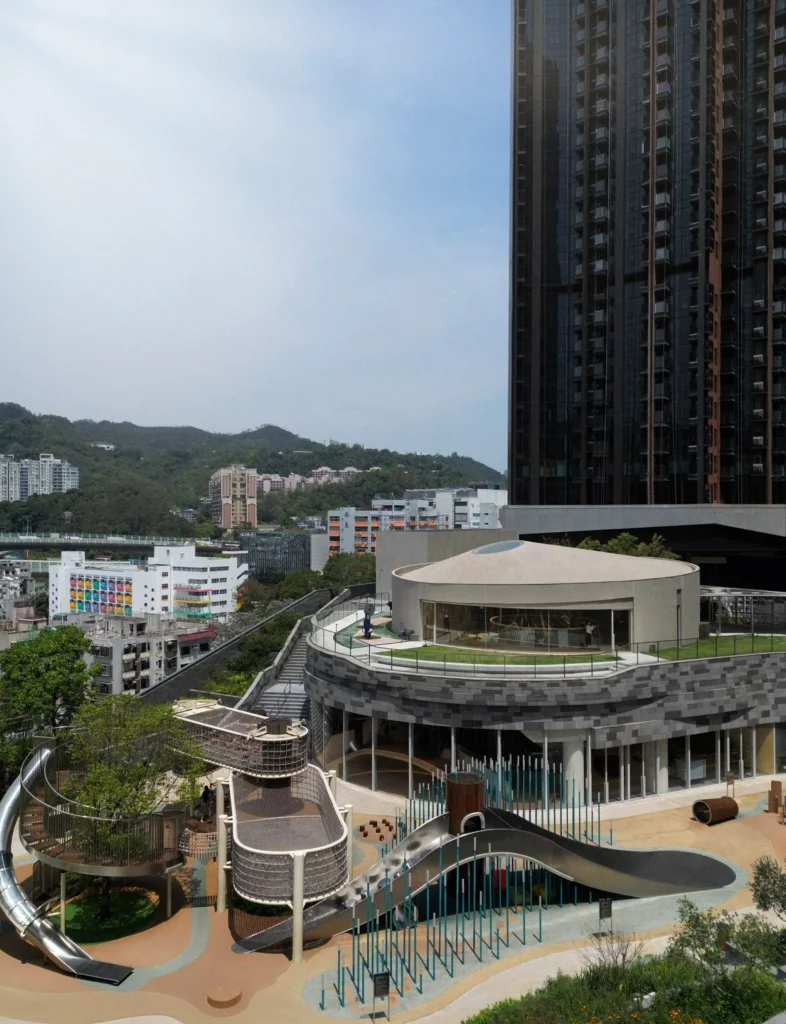 Exterior view of Sky House with outdoor playground, slides and net structures set against residential towers and green hills