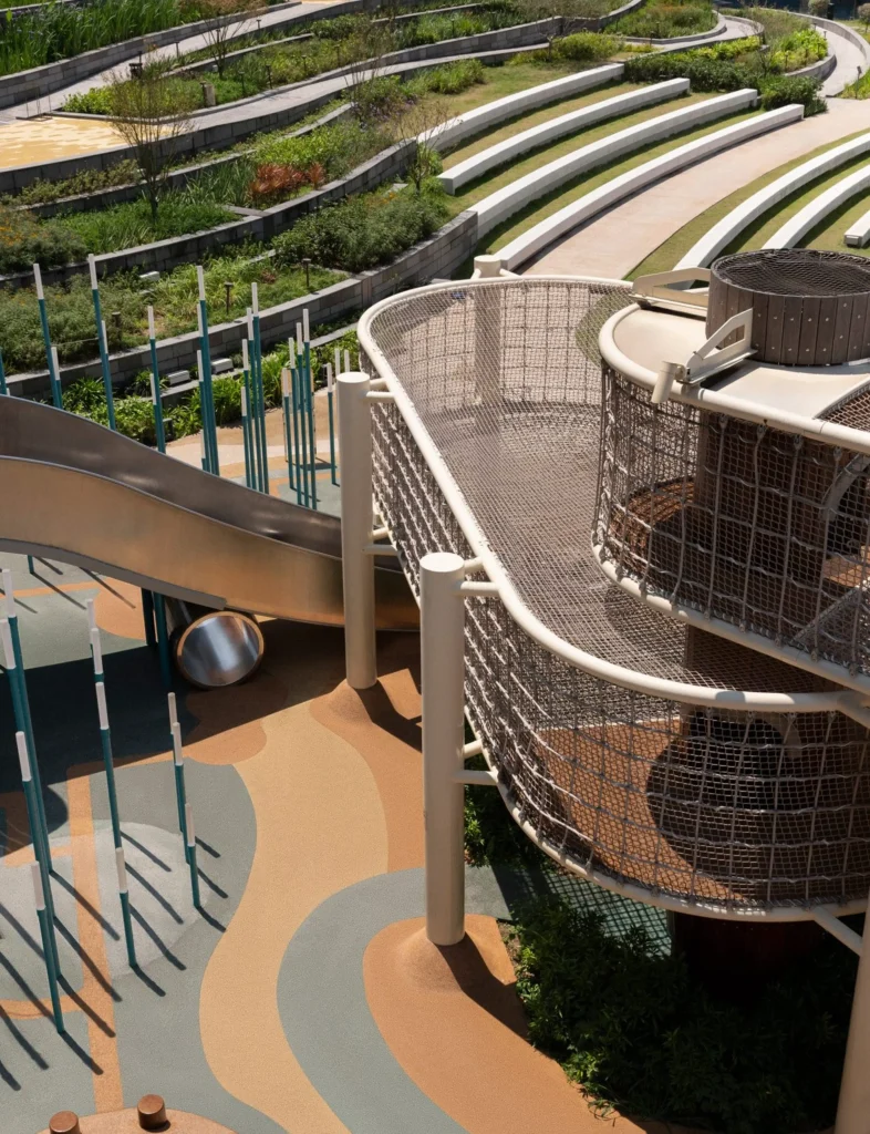 Snøhetta: Close view of outdoor playground equipment including a curved metal slide, net tower and vertical poles beside terraced planting