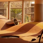 Sunlit indoor play mounds with a metal tunnel opening and views to outdoor net structures beyond