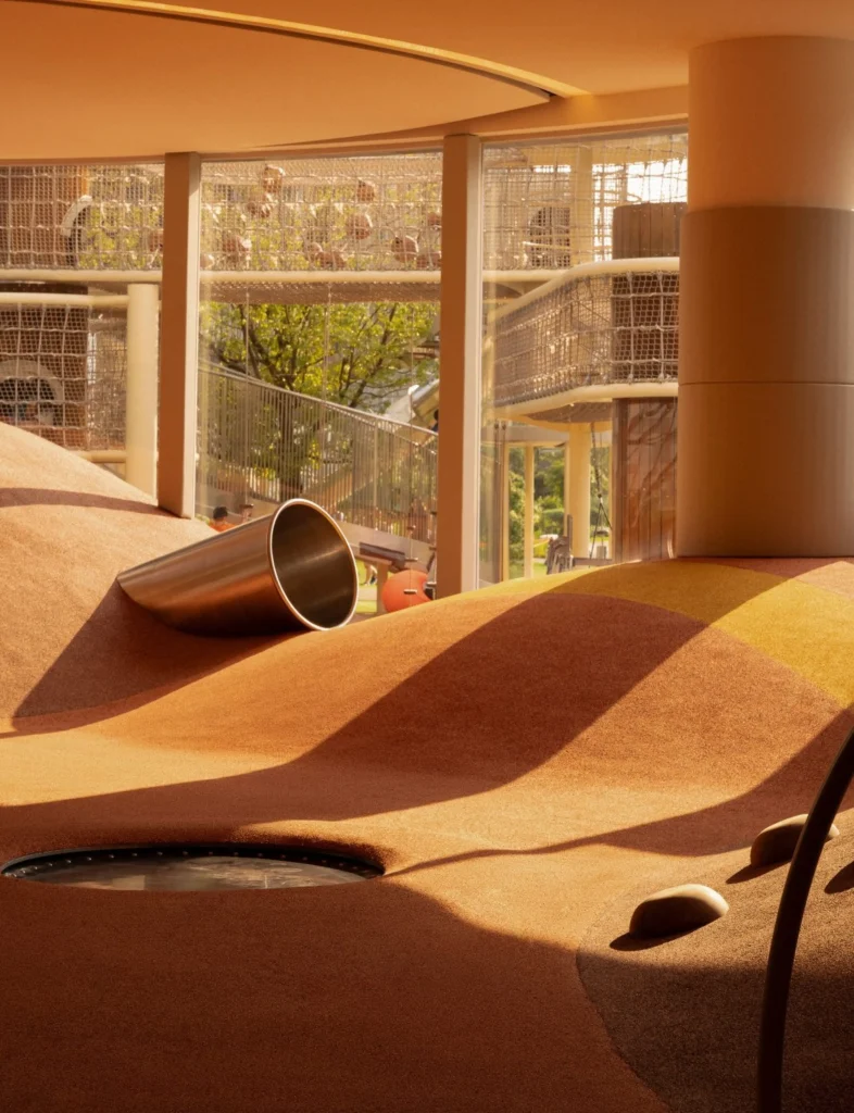 Sunlit indoor play mounds with a metal tunnel opening and views to outdoor net structures beyond
