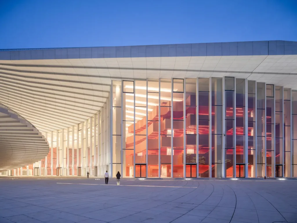 Night exterior showing the illuminated glass facade with red sculptural volumes visible inside