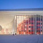 Night exterior showing the illuminated glass facade with red sculptural volumes visible inside