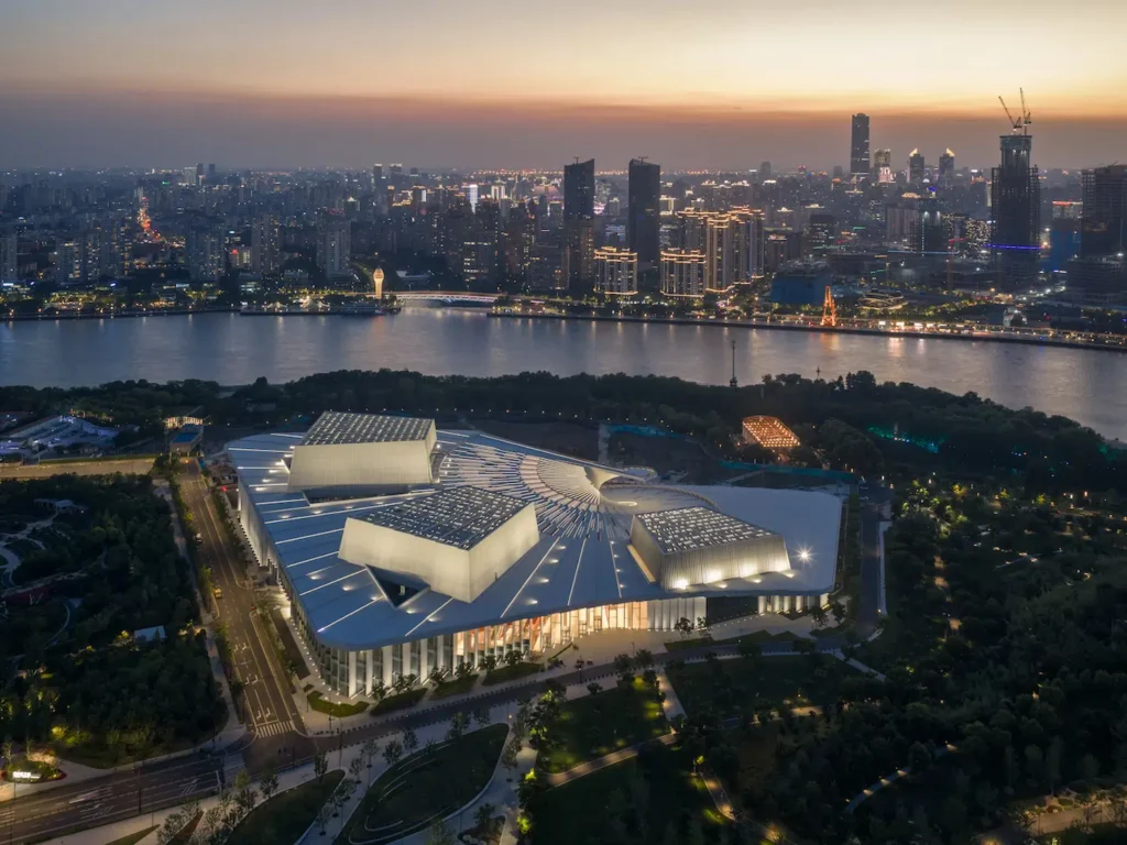 Dusk aerial of the fully lit opera house complex beside the Huangpu River and Shanghai skyline