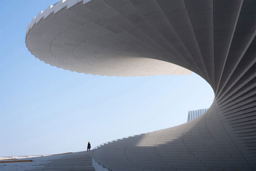 Shanghai Grand Opera House by Snøhetta Opens October 17_Low-angle view of the sweeping white helical roof with a single person walking along the curved terrace against a clear blue sky