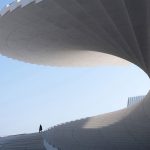 Low-angle view of the sweeping white helical roof with a single person walking along the curved terrace against a clear blue sky