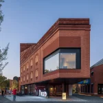 Evening view of a red brick corner building with large projecting glass window and illuminated ground floor