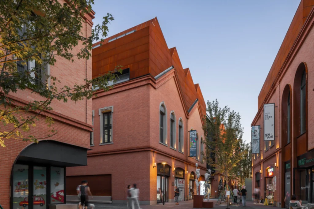 Pedestrian street lined with brick buildings, some topped with angular corten steel roofs, at dusk