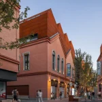 Pedestrian street lined with brick buildings, some topped with angular corten steel roofs, at dusk