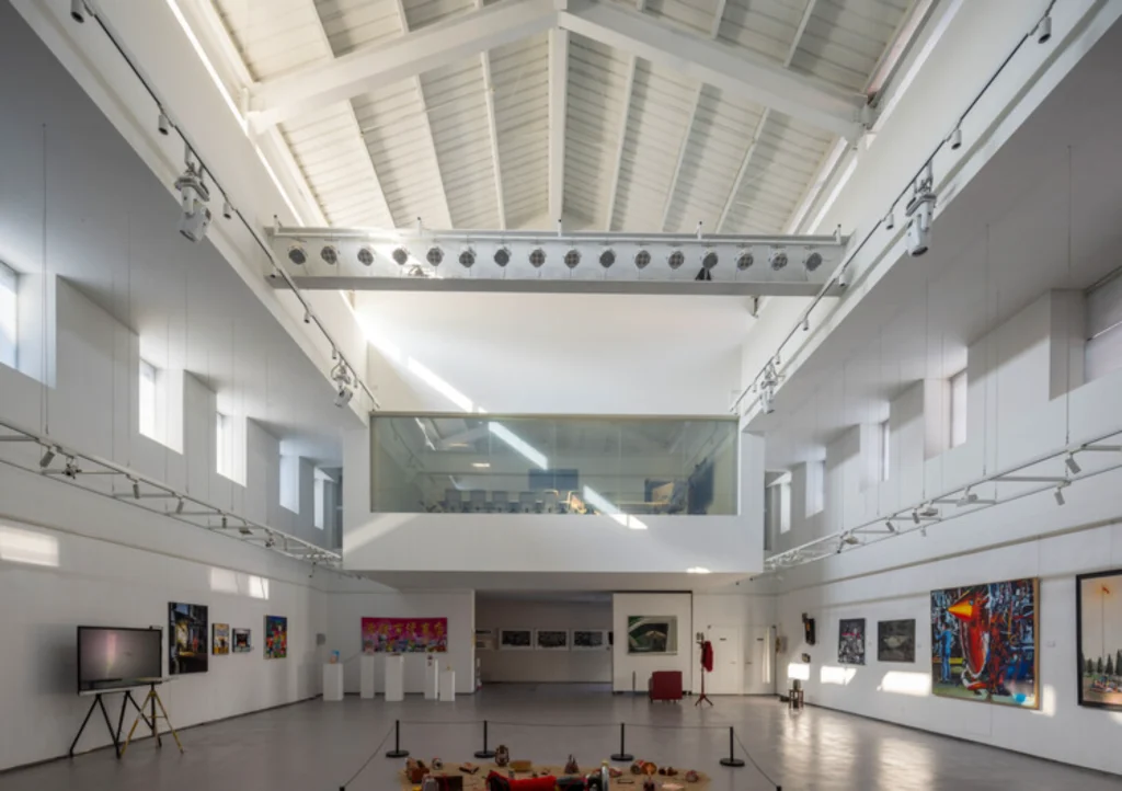 Bright double-height white gallery interior with exposed roof structure, mezzanine and artworks on display