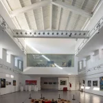 Bright double-height white gallery interior with exposed roof structure, mezzanine and artworks on display