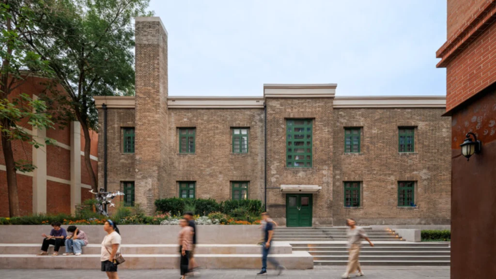 Historic grey brick building with green doors and windows facing a stepped public plaza with people