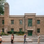Historic grey brick building with green doors and windows facing a stepped public plaza with people