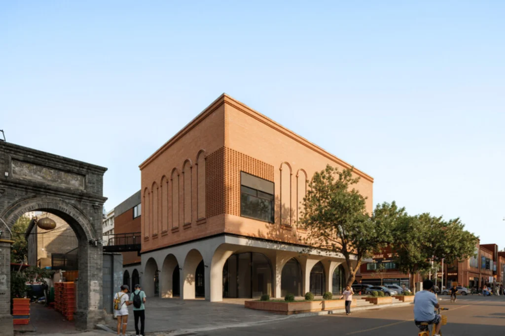 Corner view of a two-storey brick building with arched ground floor and upper arched windows beside a historic stone gate