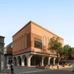 Corner view of a two-storey brick building with arched ground floor and upper arched windows beside a historic stone gate