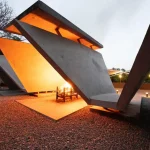 Illuminated concrete pyre at dusk with a fire burning beneath the angled planes, casting a warm glow on the gravel ground