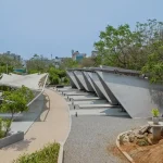 Series of angled concrete pyre structures and tensile fabric waiting canopies arranged along a curved pathway in a landscaped setting