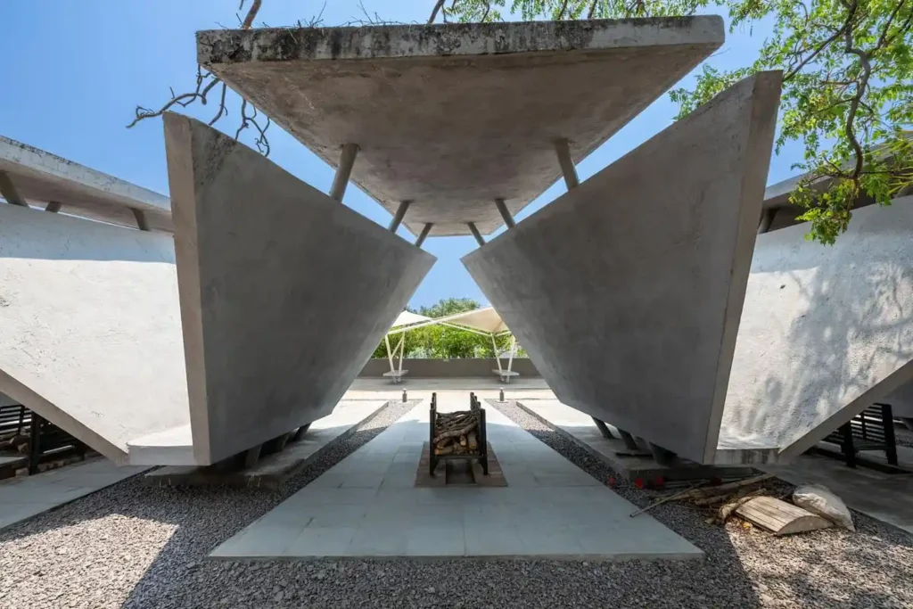 View looking through two large inclined concrete planes toward a central fire grate and distant tensile canopy
