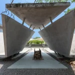 View looking through two large inclined concrete planes toward a central fire grate and distant tensile canopy