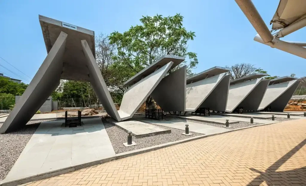 Row of angular concrete pyre structures with open sides and waiting benches, labelled Moksh Sthal under a clear sky